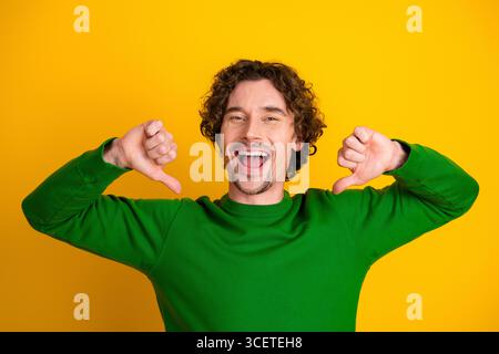 Cheerful young man in a green sweater posing enthusiastically with thumbs pointing towards himself on yellow background Stock Photo