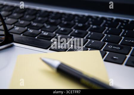 Laptop keyboard with reading glasses, pen, and yellow sticky note pad on a desk, symbolizing office work, productivity, and business planning. Stock Photo