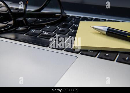 Laptop keyboard with reading glasses, pen, and yellow sticky note pad on a desk, symbolizing office work, productivity, and business planning. Stock Photo