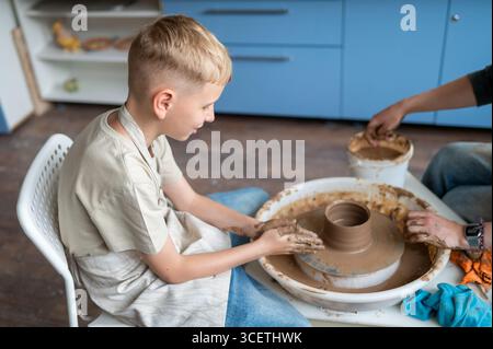 Boy and female instructor shaping clay together at pottery wheel, kids learning traditional craft Stock Photo