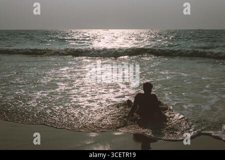 This image captures a silhouette of a person sitting at the edge of the sea, gazing at the waves and sunlight reflecting on the water, evoking a peace Stock Photo