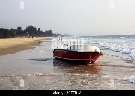 This image shows a red and white motorboat resting on the shore, with gentle waves washing around it and a distant sandy beach lined with trees Stock Photo