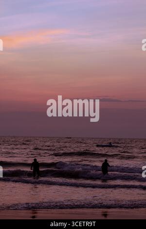 This image captures two people standing in the gentle waves of a sea or ocean at dusk, with a serene, pastel-colored sky overhead and a boat visible Stock Photo