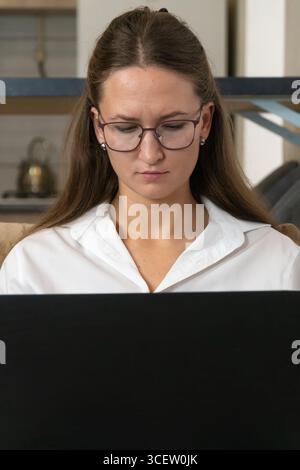 Businesswoman wearing glasses and a white shirt sits comfortably on a couch, typing on her laptop and engaging in remote work from her home office, fo Stock Photo