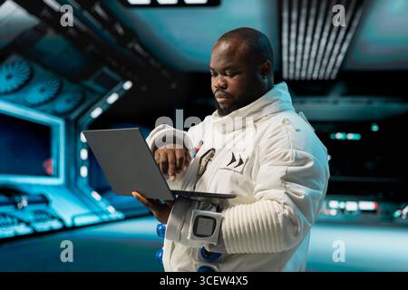 Orbit station crew member doing galaxy observation using laptop during scientific mission. Starship cosmonaut in spacesuit using notebook, monitoring system diagnostics during planetary exploration Stock Photo