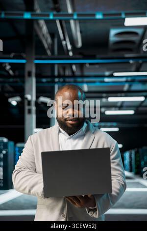 Engineer in server farm managing infrastructure systems, checking recovery plan on laptop. Data center worker monitoring performance using automation tools, making sure rigs are functioning optimally Stock Photo