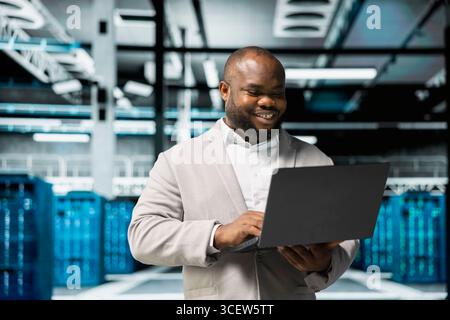 Smiling technician in data center workspace running system diagnostic tests and error checking utilities to fix problems. Happy employee looking for network issues affecting rigs performance Stock Photo