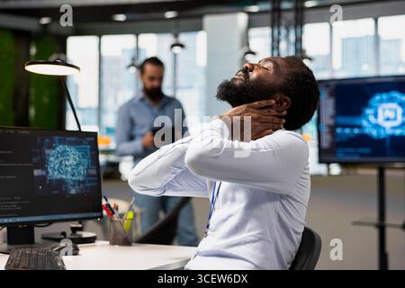 Tired engineer in office relaxing stiff neck while using AI tools to automate routine coding tasks. Fatigued IT expert massaging neck to remove pain, using artificial intelligence to do development Stock Photo