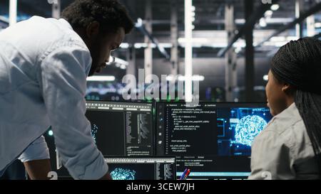 Data center engineering team uses artificial intelligence deep learning models to configure gear. Teamworking colleagues work on AI programming language in server farm, optimizing system, camera B Stock Photo