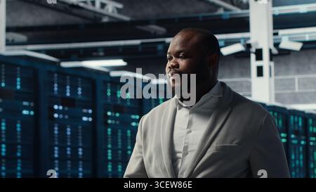 Smiling engineer in server hub monitoring data center energy consumption across components. Happy IT expert walking in data center, making sure rigs sensors are functioning optimally Stock Photo