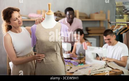 Female student works with mannequin, others pupils sewing, working in background Stock Photo