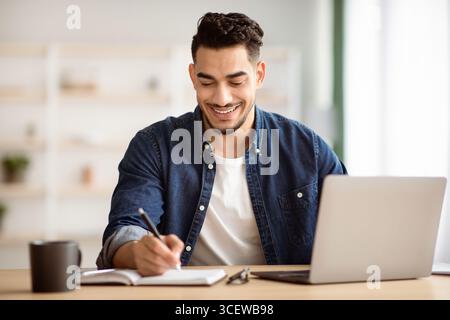 Joyful arab man taking notes while working with laptop Stock Photo