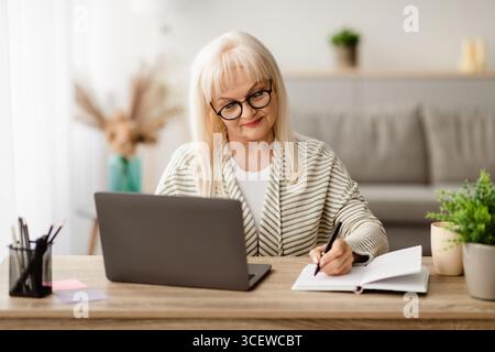 Mature lady writing in notebook and using laptop at home Stock Photo