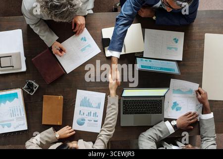 Top view showing group of middle aged multiethnic business professionals collaborating at table, two people shaking hands over laptop and financial charts, others reviewing documents Stock Photo