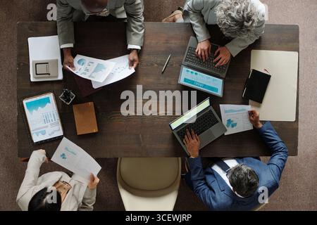 Group of middle aged multiethnic business professionals working at table analyzing financial charts and graphs using laptop and printed documents Stock Photo