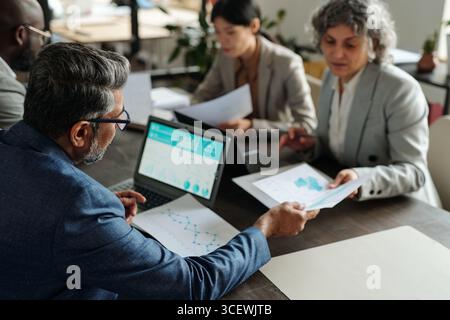 Middle aged Caucasian man analyzing financial charts on laptop while senior Caucasian woman reviewing documents with young Asian woman collaborating at office table during meeting Stock Photo