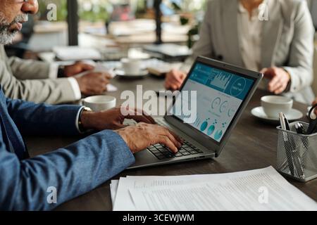 Middle aged Black man working on laptop analyzing financial charts during business meeting with two colleagues, diverse group discussing data at table Stock Photo