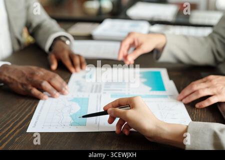 Top view showing hands of middle aged Caucasian woman, middle aged Black man, and middle aged Caucasian man analyzing financial charts and discussing business strategy at desk Stock Photo
