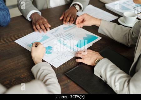 Caucasian woman and Black man discussing financial charts and graphs during business meeting, hands gesturing over printed documents on wooden table, top view composition Stock Photo
