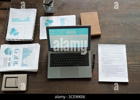 Top view showing open laptop displaying charts and graphs on screen, surrounded by printed financial reports, pen, notebook, and office supplies on wooden desk Stock Photo