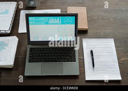 Top view showing open laptop displaying data analytics dashboard on wooden desk surrounded by printed charts, pen, documents and closed notebook, no people visible Stock Photo