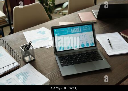 Top view showing open laptop displaying business analytics dashboard with charts and graphs on desk surrounded by printed financial reports Stock Photo