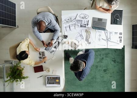 Top view of multiethnic group of young adult men and middle aged woman collaborating on architectural project, discussing blueprints and using laptop at shared workspace Stock Photo