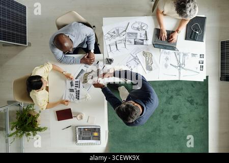 Diverse group of middle aged professionals collaborating on architectural project, reviewing blueprints and discussing plans around table, one using laptop, top view Stock Photo