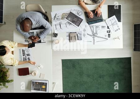 Top view showing Black middle aged man, Asian young adult woman and Caucasian young adult man collaborating on architectural project, reviewing blueprints and using laptop at workspace Stock Photo