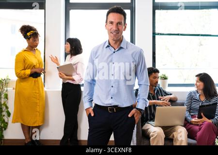 Mature businessman stands confidently with his hands in his pockets, smiling while his diverse team collaborates and works in the background of a mode Stock Photo