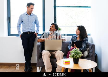 Three diverse business professionals are engaging in a collaborative discussion, utilizing a laptop and tablet, within a contemporary office startup s Stock Photo