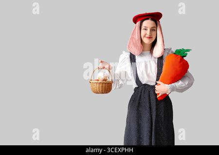 Stylish woman with bunny ears holding carrot toy and basket with Easter bread on grey background. Easter celebration Stock Photo