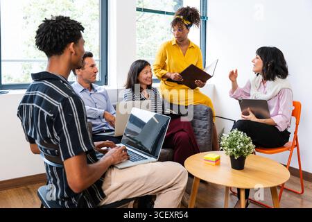 Multi-ethnic business team collaborating on project, using laptops, tablet and sharing ideas in modern office space, fostering teamwork and innovation Stock Photo