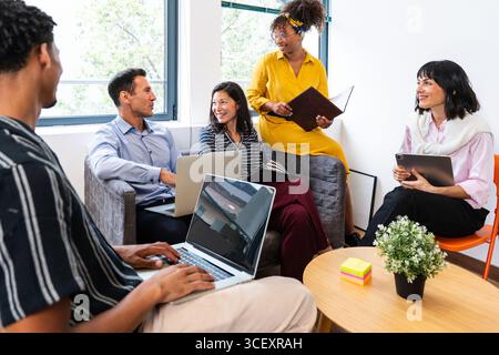Five business professionals collaborating in a bright, modern office, engaging with laptops and tablets while sharing innovative ideas and discussing Stock Photo