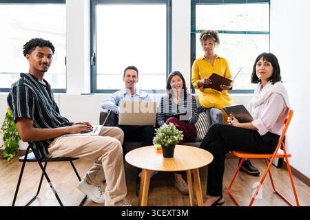 Five smiling businesspeople using laptops and tablets are having a meeting in a modern office, collaborating and working together on a startup project Stock Photo