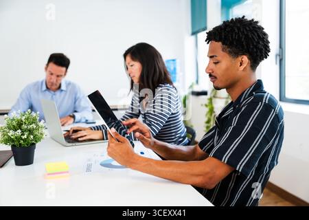 Office workers collaborating on a project at a white table, using digital devices and discussing documents, creating a dynamic and productive work env Stock Photo
