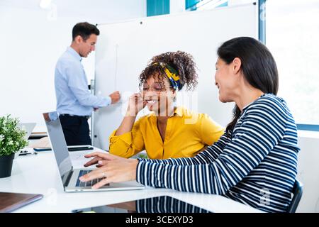 Two businesswomen are using a laptop and smiling while a businessman writes on a whiteboard during a productive meeting in a modern office, showcasing Stock Photo