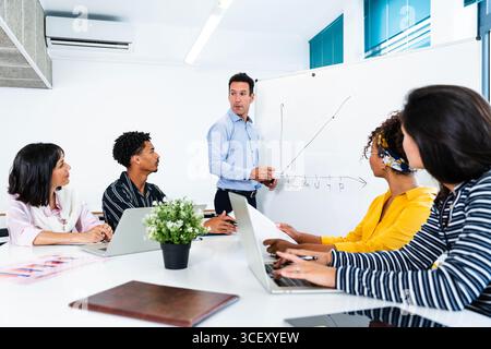 Startup business team collaborating on project during office meeting, analyzing growth chart presented on whiteboard by their manager using laptops an Stock Photo