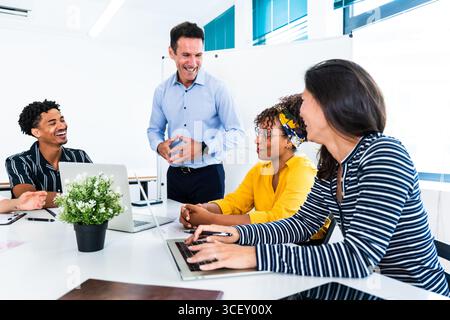 Startup business team laughing and collaborating on laptops during an engaging office meeting, showcasing teamwork and fostering a vibrant, positive w Stock Photo