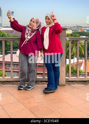 Two asian women in casual attire & headscarves taking a selfie with a smartphone on rooftop terrace with view of malacca city malaysia in background Stock Photo
