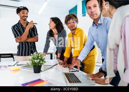 Diverse team of professionals leaning over a table, analyzing data on a laptop and documents, working together in a modern office environment during a Stock Photo