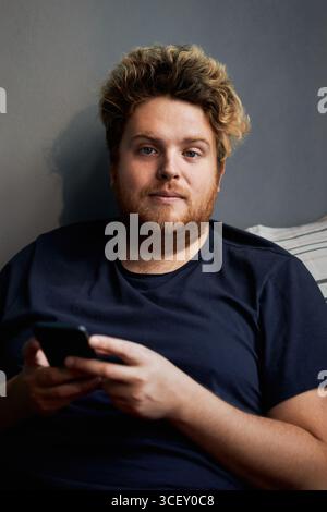 Vertical medium shot of handsome bearded Caucasian man with blond hair sitting on bed in morning holding smartphone looking at camera Stock Photo
