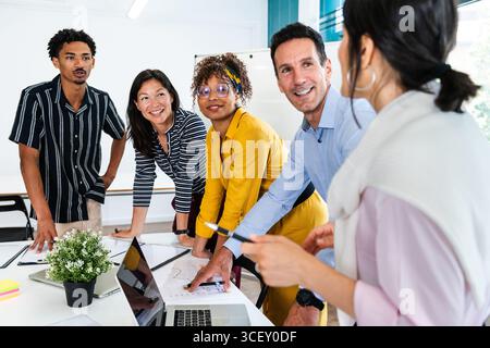 Diverse team of professionals gathered around a table, engaging in a productive discussion, analyzing charts and data on a laptop, fostering teamwork Stock Photo