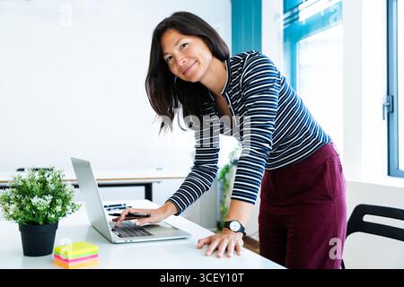 Asian businesswoman leaning over a desk, working on a laptop in a modern startup office, smiling confidently while engaging in her project and showcas Stock Photo