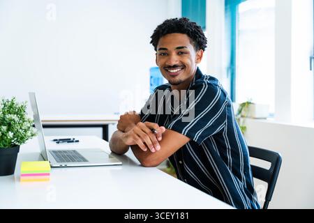 Young businessman sitting at a modern office desk, working diligently on a laptop while smiling confidently, exuding a sense of competence and profess Stock Photo
