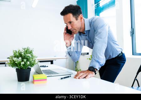 Businessman leaning on a white desk, making a phone call while working at his laptop in a modern office, with a small potted plant and sticky notes ne Stock Photo