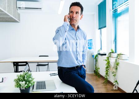 Confident businessman sitting on desk and talking on mobile phone in modern office during a working day, using laptop computer and other devices Stock Photo