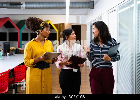 Three businesswomen are walking through a modern office, discussing work and holding digital tablets and folders, collaborating on a project in a dyna Stock Photo