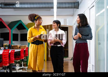 Three businesswomen are walking down an office corridor, discussing work and holding digital tablet and folders, collaborating on a project in a moder Stock Photo