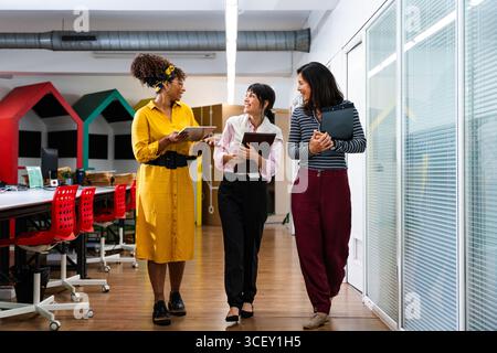 Three businesswomen are walking down an office corridor, holding digital tablets and clipboards, discussing work and smiling, showing teamwork and col Stock Photo